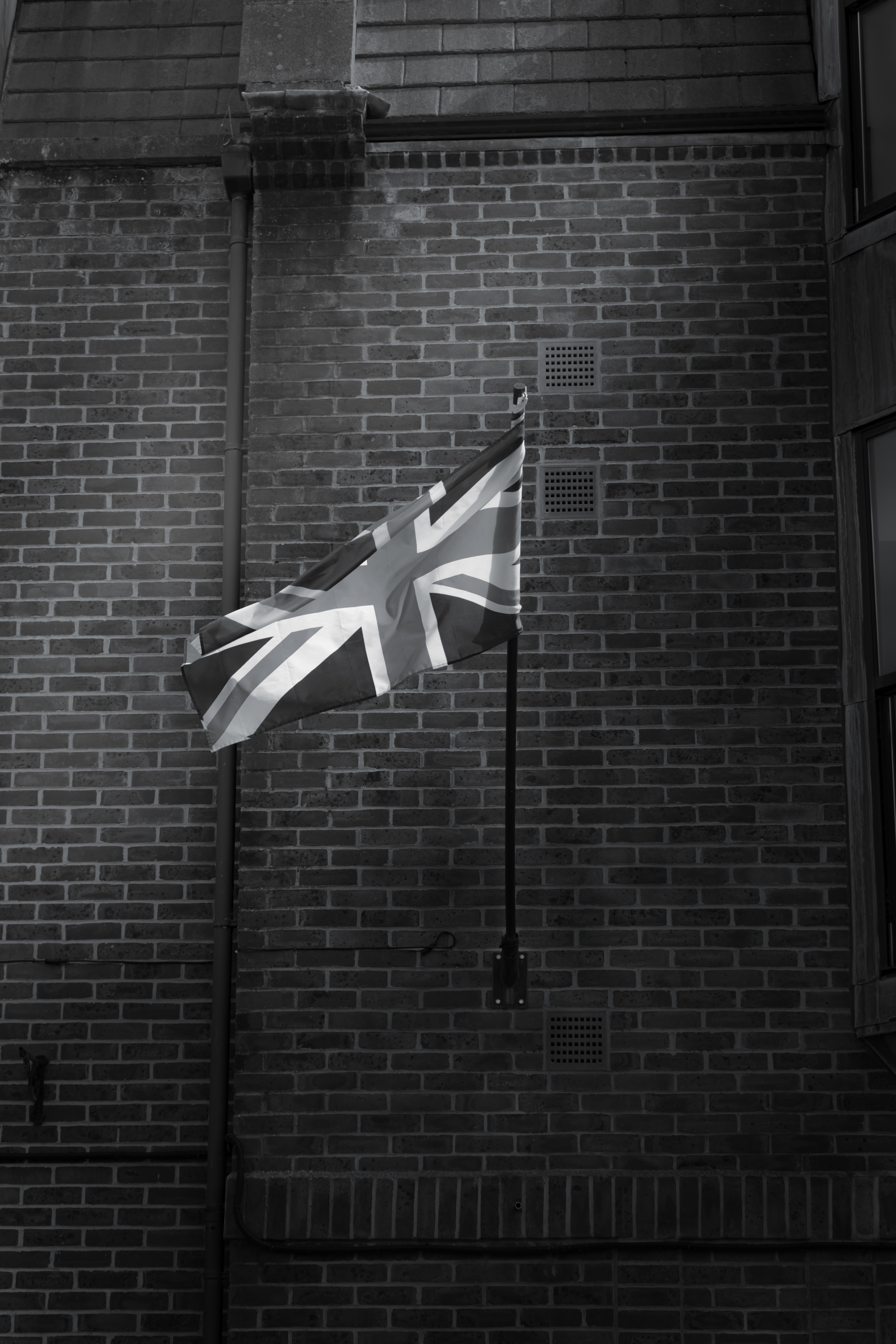 A dramatic black and white photograph of the Union Jack flag which is erectred from a pole connected to the wall of a brick building.